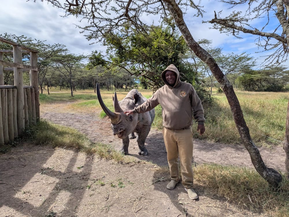 Our founder standing relaxed beside a black rhino under acacia trees, one hand resting on its back.