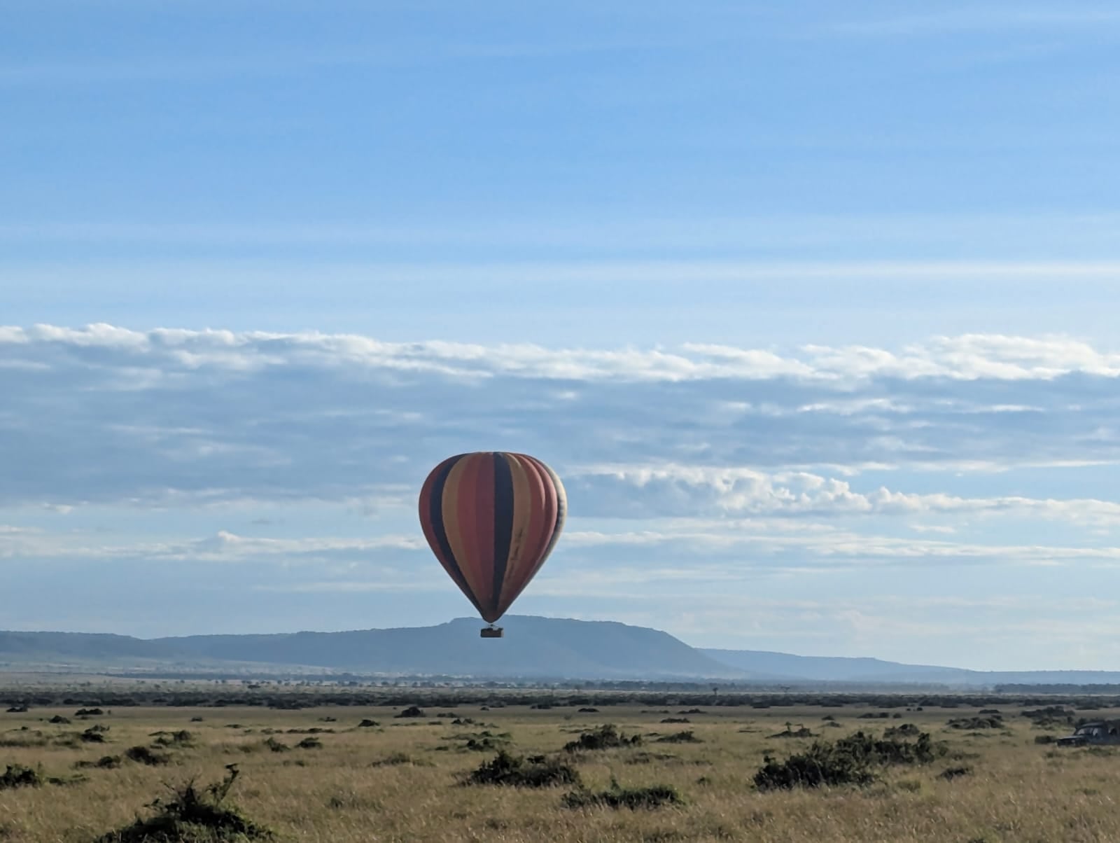 A single hot-air balloon hanging low over the grass plains of the Maasai Mara, with the escarpment on the horizon under a wide pale sky.